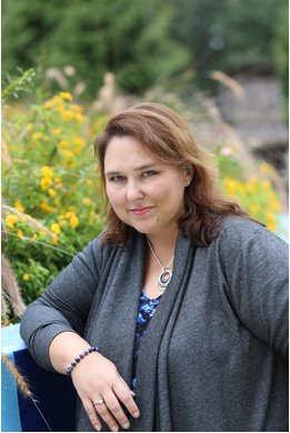 Jennifer Burke Mighty Marketing Mojo marketing coach brunette in grey knit top leaning on wall smiles at camera